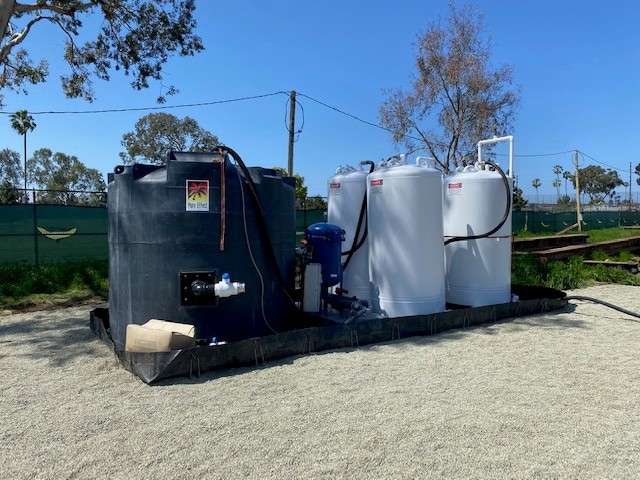 An installed Pure Effect groundwater treatment train with a black poly tank and two white carbon vessels on an outdoor secondary-containment berm